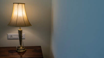 An electric lamp on a wooden bedside table in the corner of the room. Metal decorative stand and translucent elegant lampshade. The lights are on.