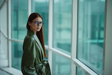 Fashion woman in a stylish raincoat and sunglasses standing by the window and looking at the city...