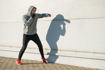 A man in a hoody trains boxing against a gray wall. 