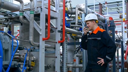 A young inexperienced engineer at his workplace in a white helmet and overalls inspects the equipment of the compressor station. Work at an industrial plant in an industrial workshop.