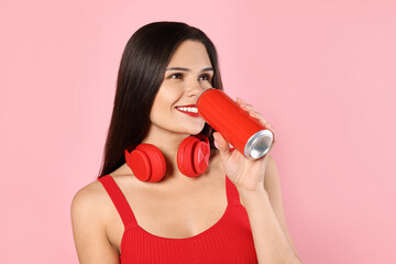 Beautiful young woman with headphones drinking from tin can on pink background