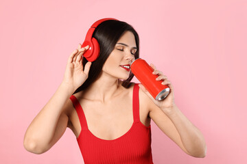 Beautiful young woman with headphones drinking from tin can on pink background