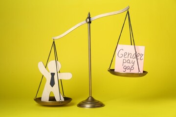 Gender pay gap. Wooden figure of man and paper note on scales against yellow background