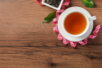 Cup of herbal diet tea and measuring tape on wooden table, flat lay with space for text. Weight loss concept