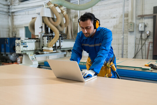 Male carpenter worker working with laptop computer in the wood workshop. Male worker in safety uniform for control CNC machine at wood industry factory. Industry manufacturing concept