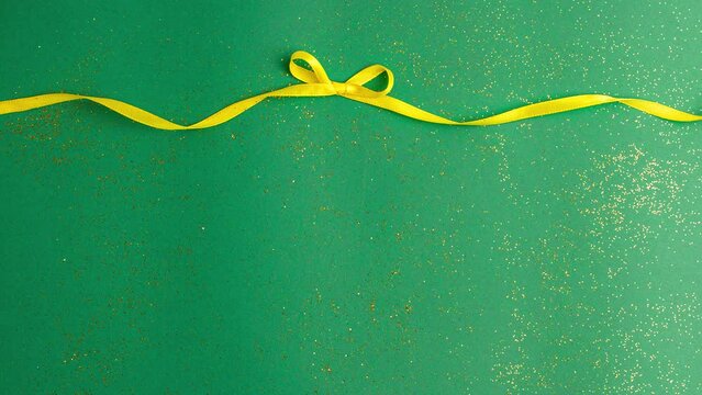A yellow satin ribbon bow tied and untied on jade green background with gold colored glitter. Stop motion animation Christmas Holidays and present concept flat lay with copy space