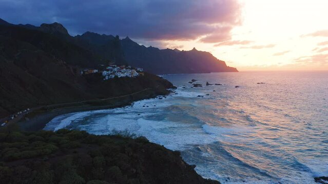 Aerial sunset light rural landscape small town surrounded by ocean waters with mountains silhouette and dark cloudy night sky. Almaciga Tenerife Canary Island Spain.