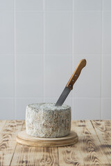 Knife in whole round blue cheese wheel on wooden cutting board on light background, selective focus