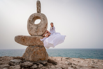 A woman sits on a stone sculpture made of large stones. She is dressed in a white long dress,...
