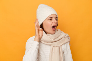Curious woman wearing warm clothes, keeps hand near ear tries to hear rumors, has intrigued expression, tries to hear conversation, posing isolated over yellow background.