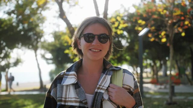 Joyful woman with short hair and sunglasses smiling outdoor looking at camera standing in city park.
