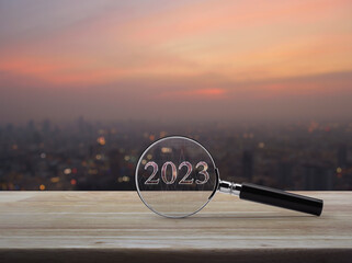 Magnifying glass with 2023 text and financial graph on wooden table over blur of cityscape on warm...