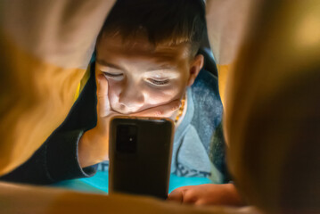 Fototapeta na wymiar Serious boy,face under the blanket at night in his bed communicates on Internet.Child gadget addiction and insomnia.Selective focus.