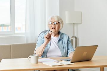 Happy smiling caucasian blonde senior woman sitting at dining table, having video call over at laptop.