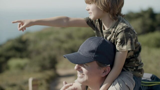 Closeup of father carrying son on shoulders on hiking adventure. Dad and child smiling, enjoying vacation together. Boy pointing at something in distance. Family, happiness, travel concept.