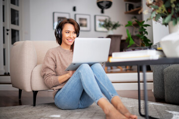 Fototapeta na wymiar Attractive and cheerful woman using laptop and headphone while sitting on the floor at home