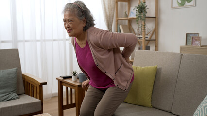Asian grandma suffering chronic backache is dropping down in the couch and massaging her lower back...