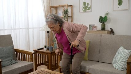 asian grey haired mature woman walking with a crutch due to injury and sitting down in the sofa feeling great discomfort in her lower back at home