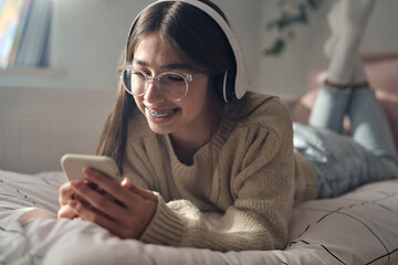 Caucasian teenage girl browsing phone with smile and wearing headphones while lying on bed