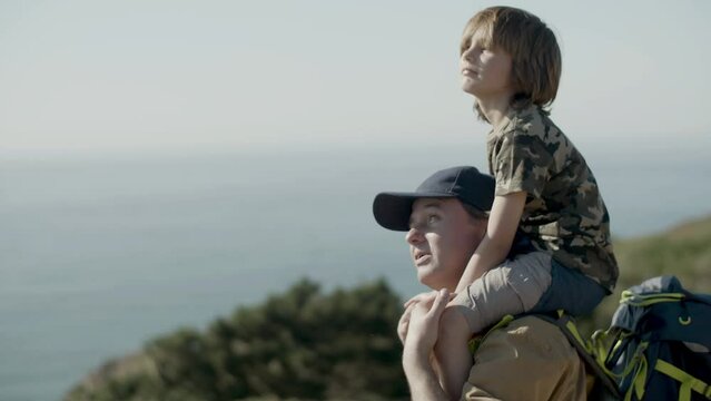 Father carrying his son on shoulders while hiking at seashore. Dad showing his boy something in distance, looking at sea and pointing. Tracking medium shot. Family, father and son, travel concept.
