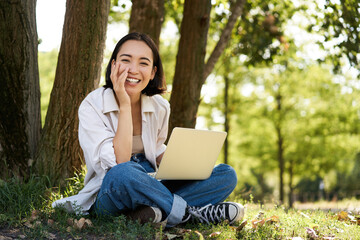 Portrait of young asian woman sitting in park near tree, working on laptop, using computer outdoors