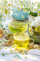 Chamomile herbal tea in a glass cup on wicker coaster on wooden table, healthy chamomile flower drink with honey, fresh chamomile flowers in background, healthcare and healthy eating concept