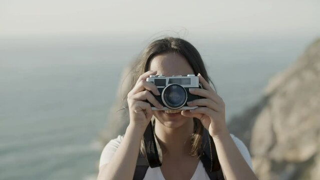 Closeup of young dark-haired woman taking photo in mountains. Smiling Caucasian girl holding camera, taking picture against picturesque sea background. Front view. Photography, travel concept.