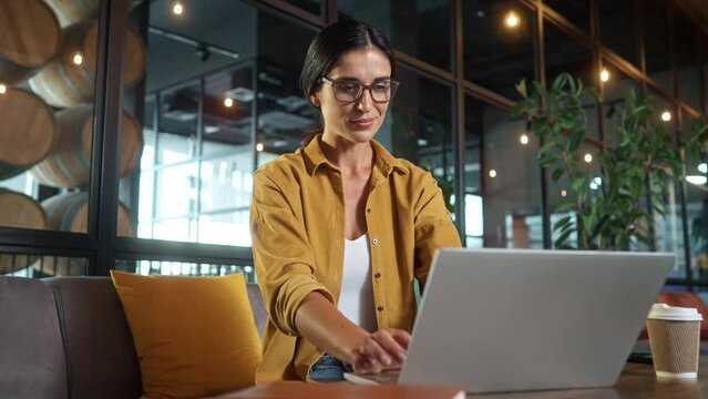 Portrait of young inspired pretty woman typing on laptop sitting on sofa in cafe working online. Successful happy female typing on computer doing tasks smiling wearing glasses. Freelance concept.