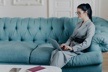 Fototapeta na wymiar Content businesswoman works remotely in cozy home office, poses with laptop computer on sofa, wears formal elegant clothes, keyboards and searches necessary information in internet, sends emails