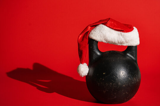 Round black kettlebell with a handle with Santa Claus hat on a red background with a shadow. Old heavy metal kettlebell for sports, bodybuilding, weightlifting or crossfit. Christmas concept.