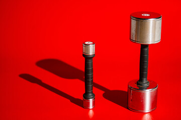 Two metal silvery dumbbells standing on red background with shadows. Small and large dumbbells of different weights for pumping up muscles. Sports equipment for fitness, crossfit, bodybuilding.