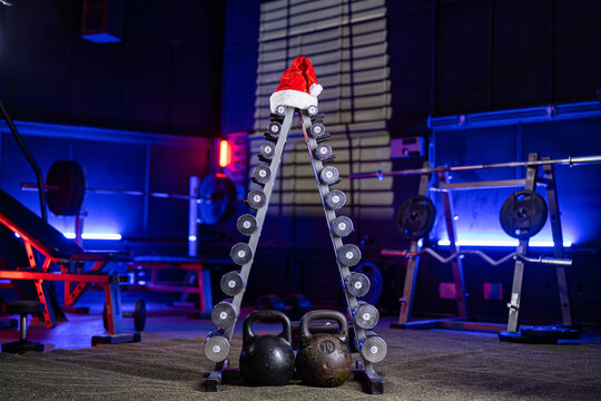 Rack with set of dumbbells in form of Christmas tree and red Santa Claus hat. Sports equipment in gym in blue light. Dumbbells, weights, barbells and sports bench for crossfit, bodybuilding, fitness.