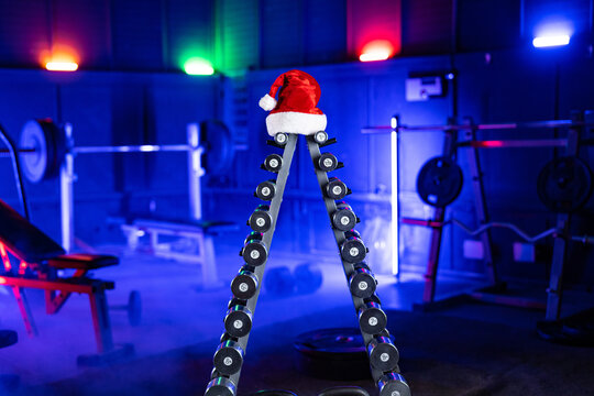 Rack with set of metal dumbbells in form of Christmas tree and red Santa Claus hat. Sports equipment in gym in blue light. Dumbbells, barbells and sports bench for crossfit, bodybuilding, fitness.
