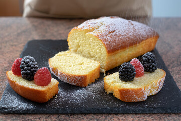 Close-up on the homemade plumcake resting on a black stone cutting board while being dusted with icing sugar. Cooking at home. Delicious healthy homemade dessert served with berries