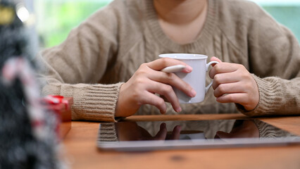 Asian woman in cozy sweater sipping morning coffee while using her digital table at desk.