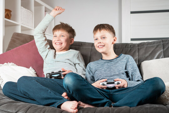 Portrait of two smiling pleased teenage boys children sitting on sofa at home, holding gaming controller joystick, playing videogames, raising hand, gesturing. Hobby, free time, gaming, friendship.