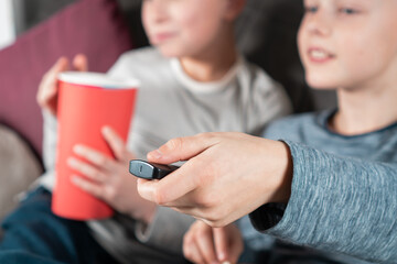 Cropped photo of two smiling teenage boys sitting on sofa at home, watching exciting movie comedy video cartoon on TV, holding remote control, eating popcorn from red bucket, having fun. Soft focus.