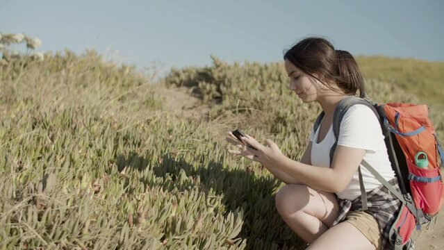 Pretty Caucasian girl with backpack taking photo of flowers. Young dark-haired woman making picture of rare plants with smartphone while hiking on sunny day. Modern technology, nature, travel concept.