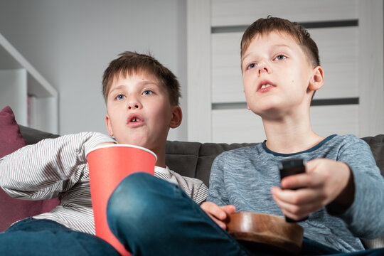 Portrait of two teenage boys sitting on sofa at home, watching interesting film movie video cartoon on TV, holding remote control, eating popcorn from red bucket and wooden bowl. Hobby, free time.