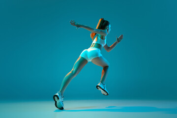 Fototapeta na wymiar Young sportive girl in white sportswear training, running isolated over blue studio background in neon light