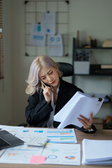 Portrait of a young Asian woman showing a serious face as she uses her phone, financial documents and computer laptop on her desk in the early morning hours