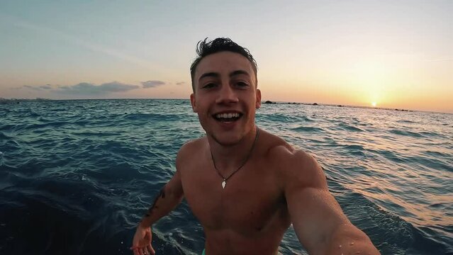One young happy man enjoying and having fun alone at the beach in the water jumping looking at the camera while taking a selfie.