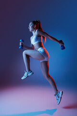 Young sportive girl in white sportswear training with dumbbells isolated over gradient blue purple studio background in neon light. High jump