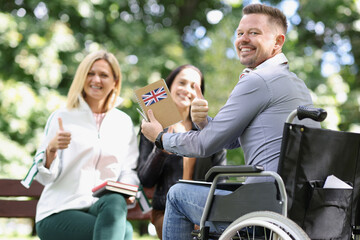 Smiling disabled man in wheelchair studying english with friends and showing thumbup sign