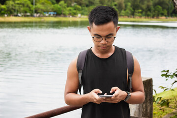 Filipino man with eyeglasses sending text message on cellphone with lake on background. Asian male using phone outdoors