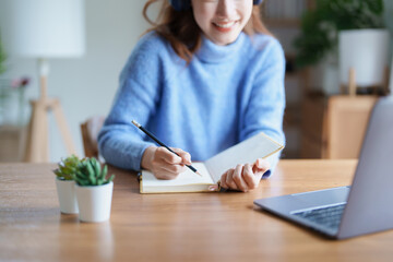 Portrait of a teenage Asian woman using a computer, wearing headphones and using a notebook to study online via video conferencing on a wooden desk in library