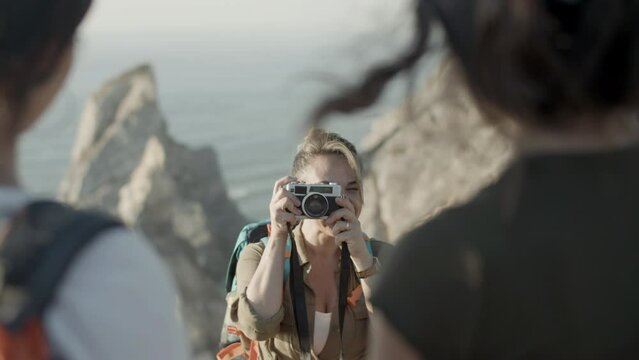 Smiling woman taking photo of her family standing on cliff edge. Caucasian blonde woman holding camera, taking picture of two people. Blurred silhouettes in foreground. Love, family, travel concept.