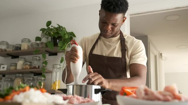 Young african man using a blender to prepare chicken mince in a kitchen.