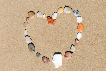 Fototapeta na wymiar Multi-colored stones laid out in the shape of a heart on the sandy seashore. Romance and relaxation. Close-up.