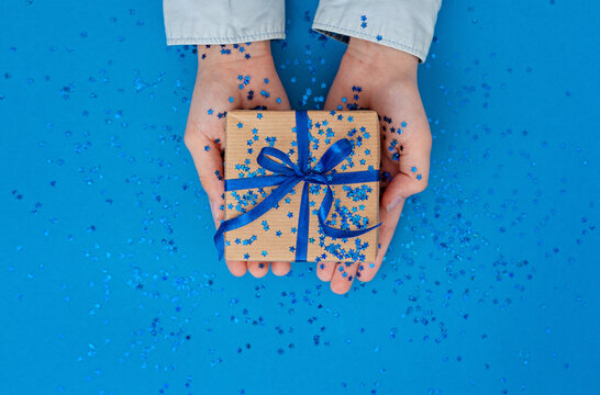 Hand giving a gift box with blue ribbon on blue paper background. Birthday or christmas holiday concept.
